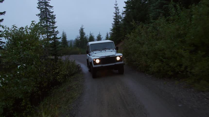 A jeep is traveling on a forest road with the headlights on