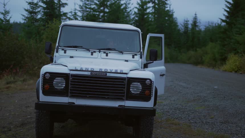 Man with grey beanie and coat opens white land rovers door while it rains in the countryside