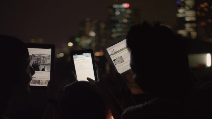 People are silhouetted against a city backdrop as they hold electronics and look at the screens
