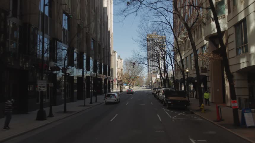 Car drives down city street lined with parked cars and leafless trees in shadow between block of buildings as pedestrians move along sidewalks