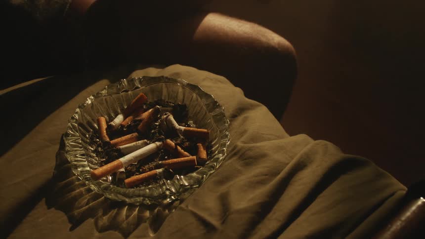 Shadow of a person sitting next to an ashtray filled with cigarettes inside a room at evening