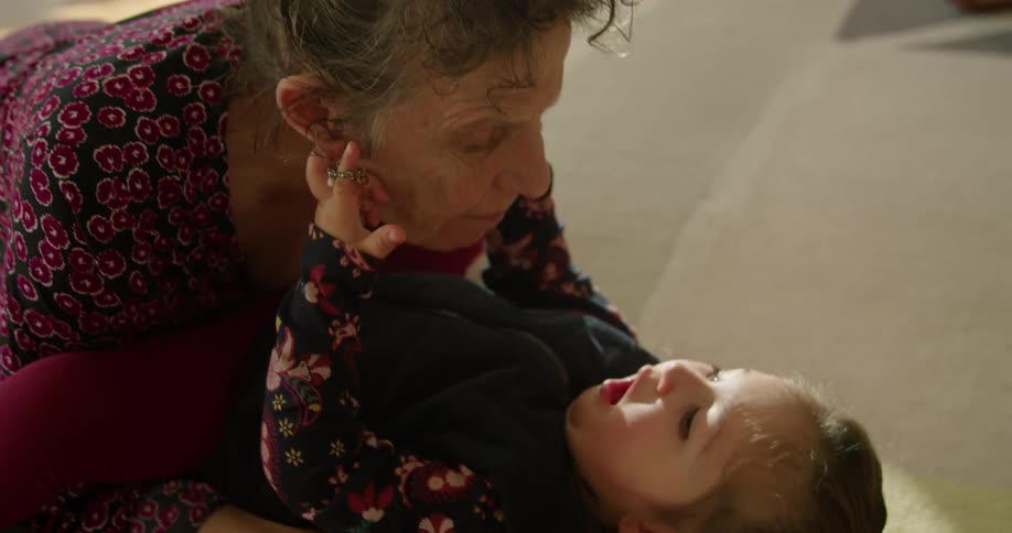A young girl lying on the carpet plays with the earrings in the ears of her grandmother who is leaning over her