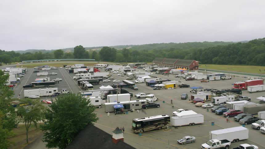People walk around parking lot among rv trailers and cars with forest on horizon under overcast sky