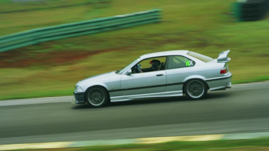Silver car races on a racetrack next to a field as trees sit in the distance