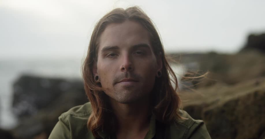 Young long haired man sitting on the rocks on a beach looking at the camera