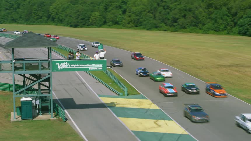Cars race around a track as they pass a tower where someone waves a flag