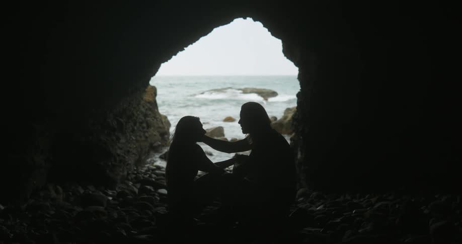A man and woman sit facing one another in a cave on the coast while caressing each others face