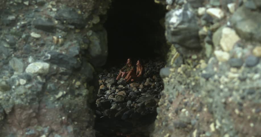 Man with long hair and woman with brown hair sits on stones in dark cave talking to each other