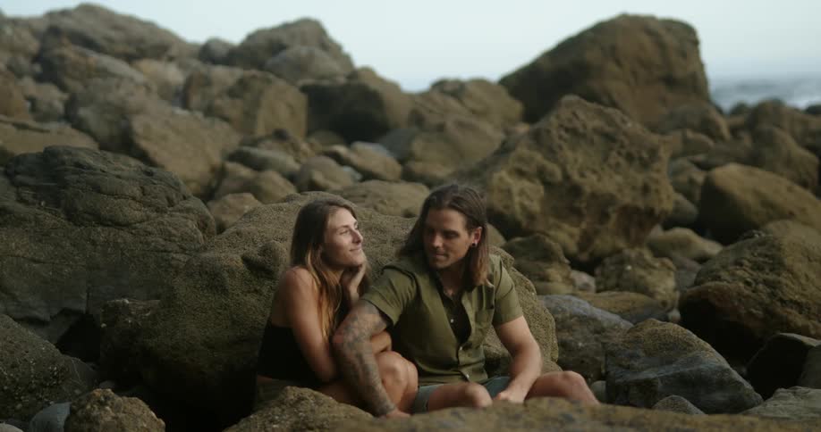 A man and the woman sitting on rocks with the sky and clouds and the ocean in the background