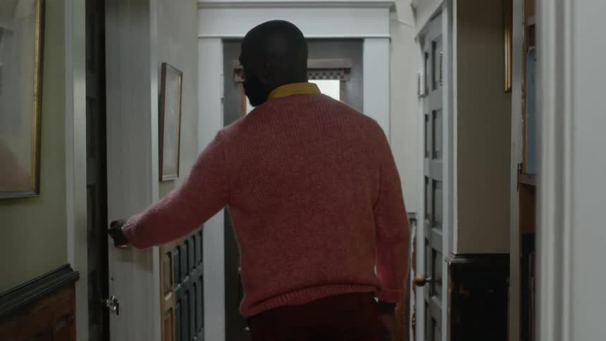 A man slowly walks down a hallway to a door that he gingerly opens