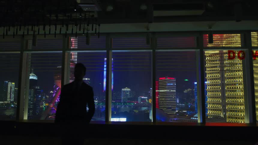 Man silhouetted by city lights walks away from windows showing city at night