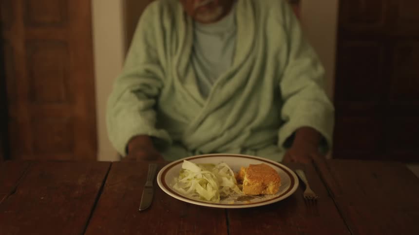 An old man in a robe sitting at the table with a plate of food
