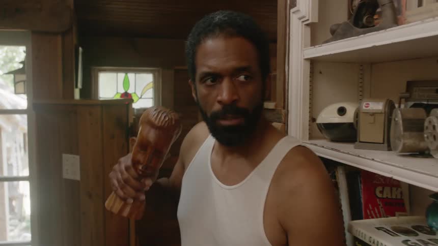 A man in a room with a wooden sculpture in his hand with a wall a shelf and door in the background