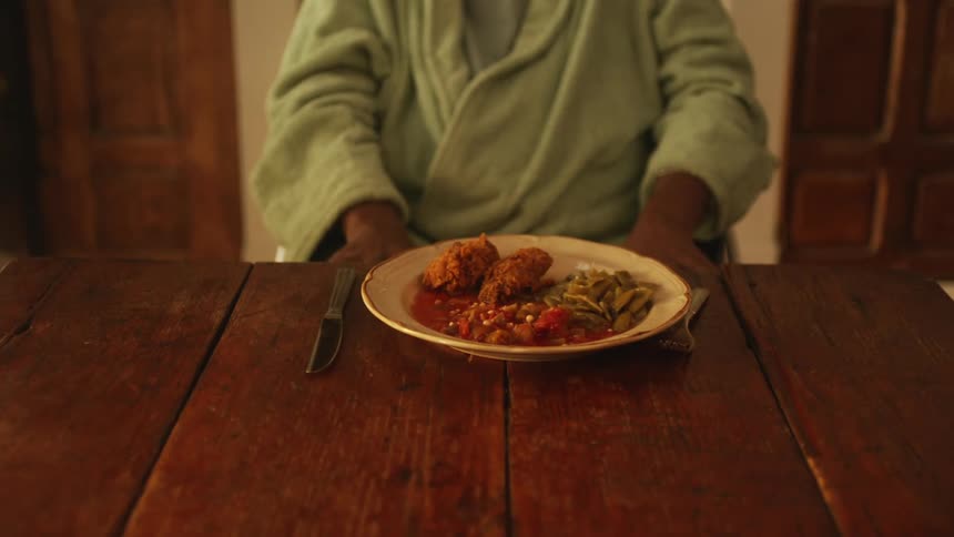 Person wearing yellow walks into room and places plate of food in front of person sitting at wooden table