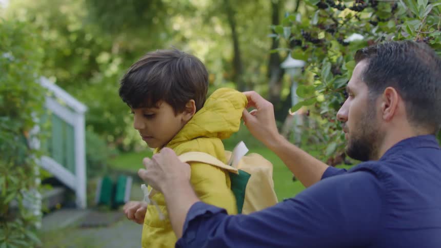 A man helps a young boy zip his jacket and put on a backpack in a backyard