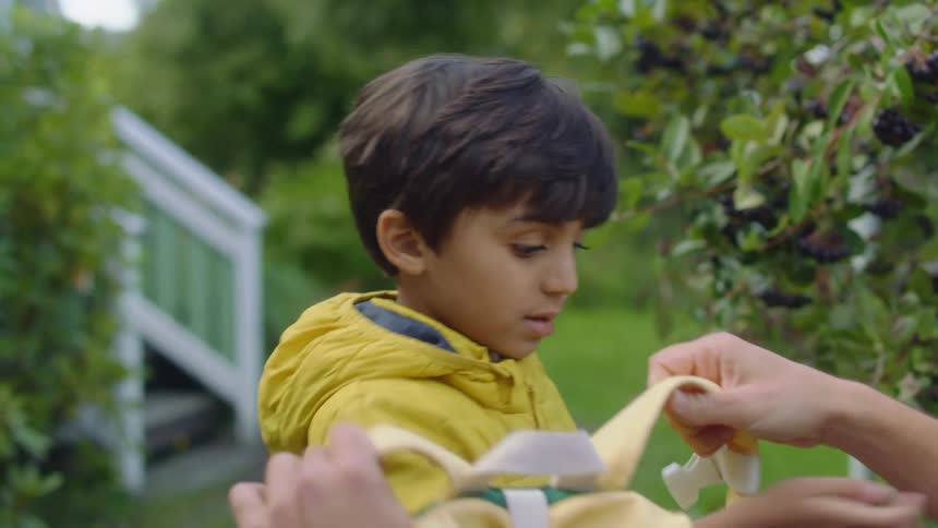A young boy zips up his jacket and his father helps him put on a backpack