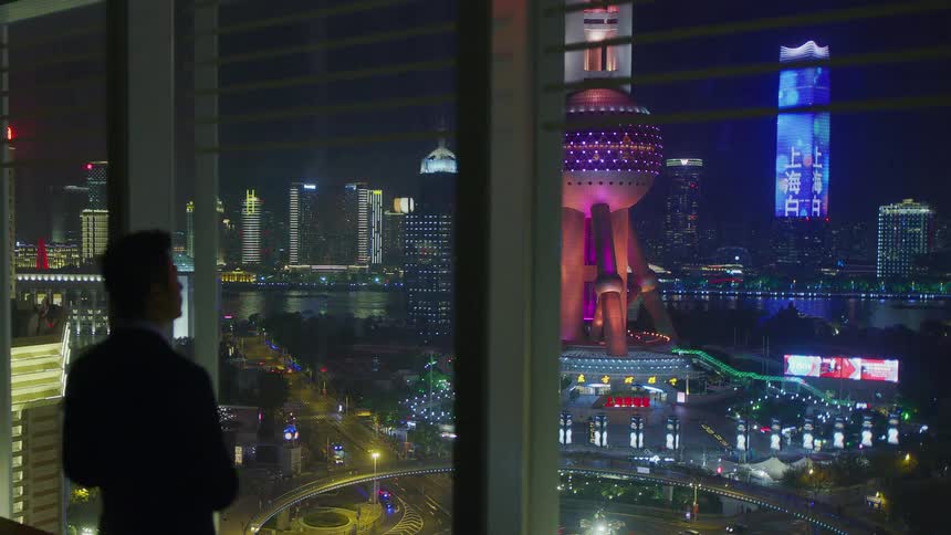A man stands in front of large building windows looking at the city skyline at night