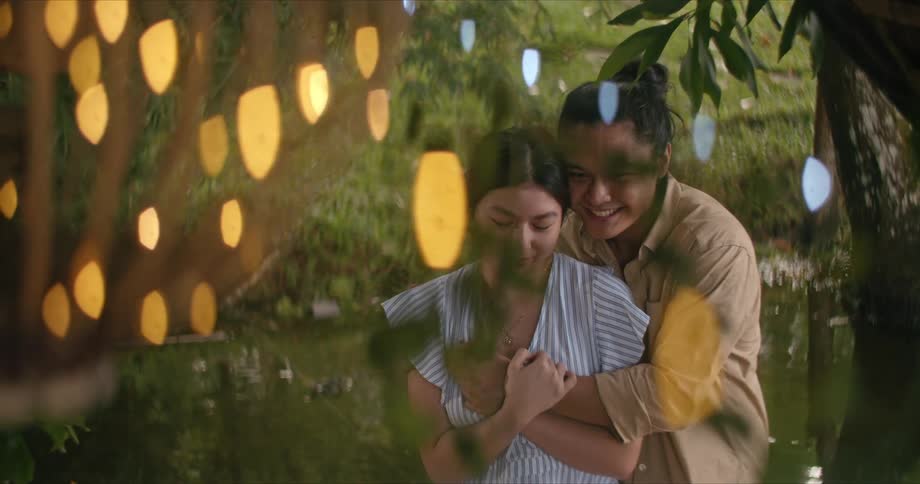 A woman and a man stand together hugging while looking at lights on the trees