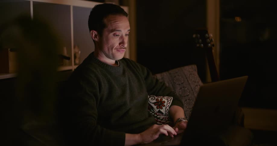 A man in a room sitting on a couch typing on a laptop with a guitar and a wall in the background