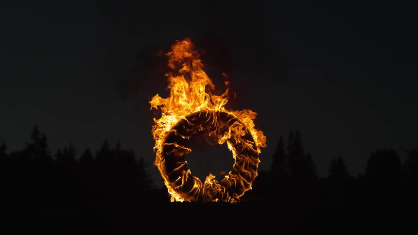 Large ring sits on end covered in orange flames releasing smoke with conifer forest silhouetted in background