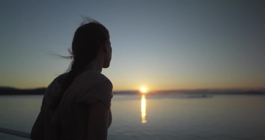 Woman leans against rail staring over ocean to sun on the red tinted horizon