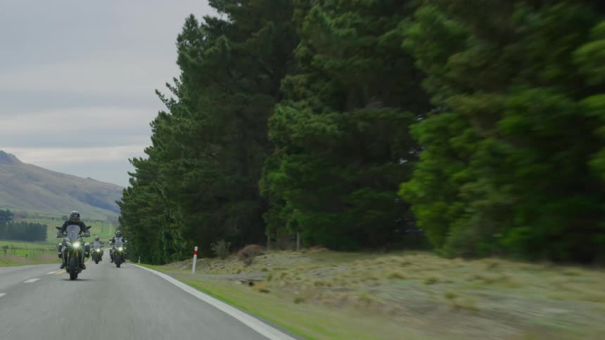 Motorcyclists ride past driveway passing conifer trees under overcast sky with mountain in background