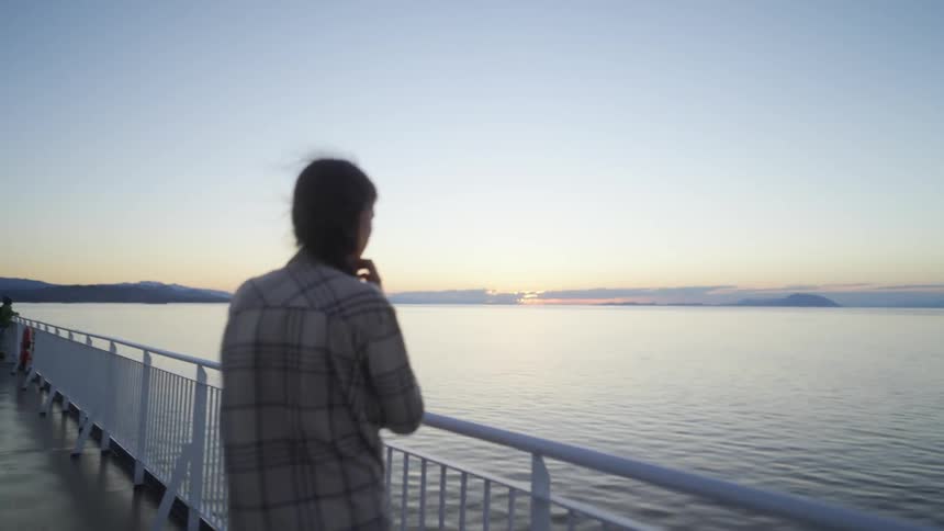 A woman walks to the railing on a ship to look out at the sunset