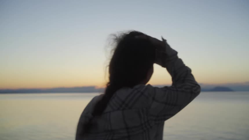 A woman brushes back her hair with her hand as she looks at the sunset on the water