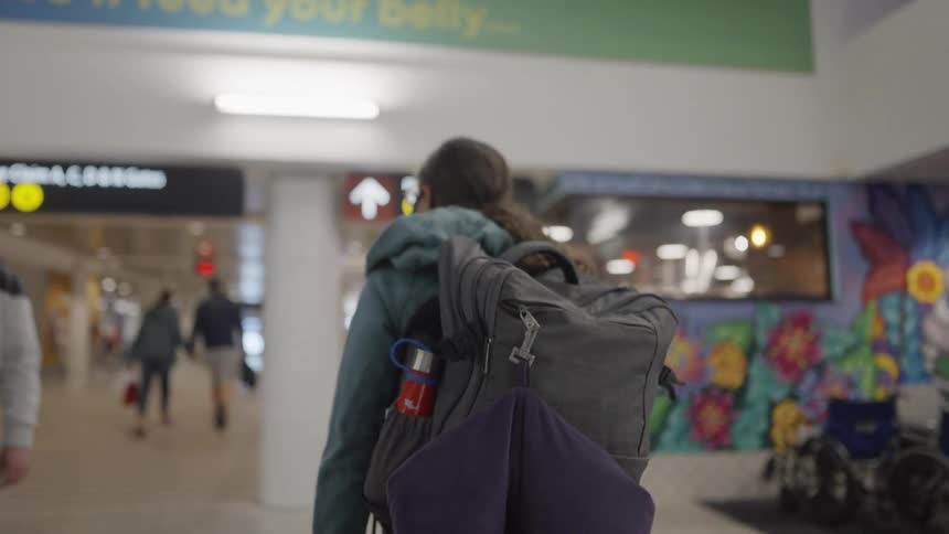 Woman wearing backpack and coat walks through airport past wall mural