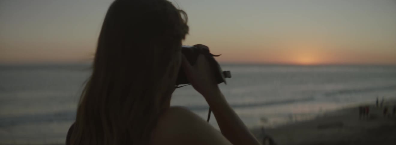A woman standing on a beach at sunset raises a camera to her eyes