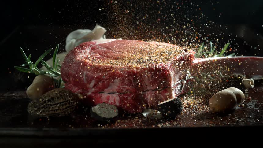 A heavy amount of seasoning is poured onto a raw tomahawk steak over a plate of vegetables and herbs