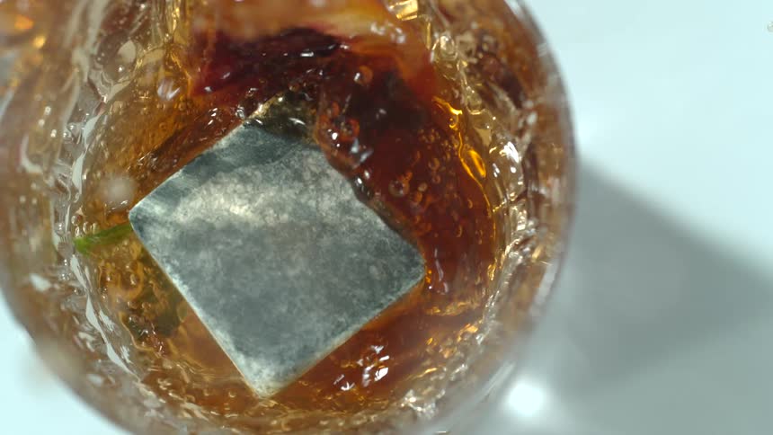 A gray cube drops into a cocktail glass splashing amber liquid up and out of the garnished glass