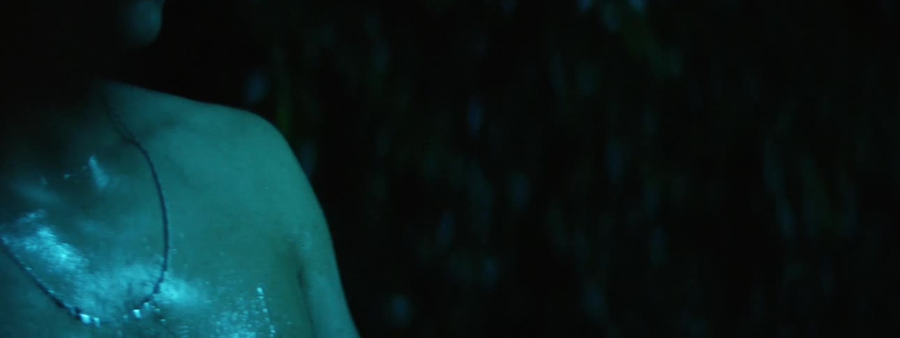Bare chested sweating man stands outdoors near vegetation under blue colored lights