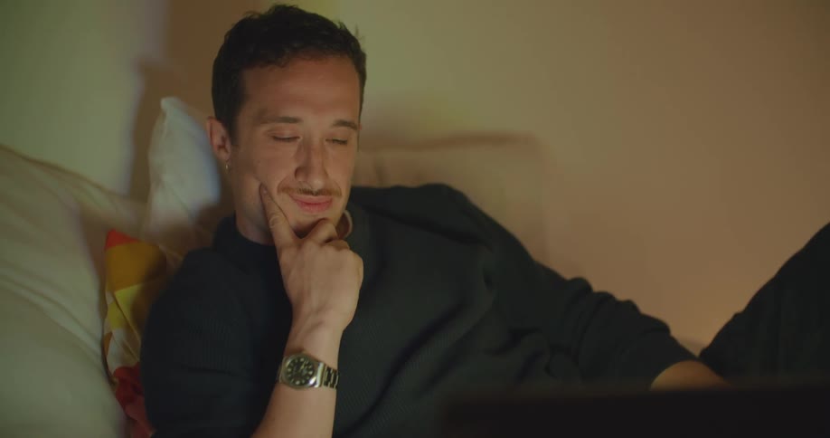 A man smiles as he lounges in bed looking at his laptop