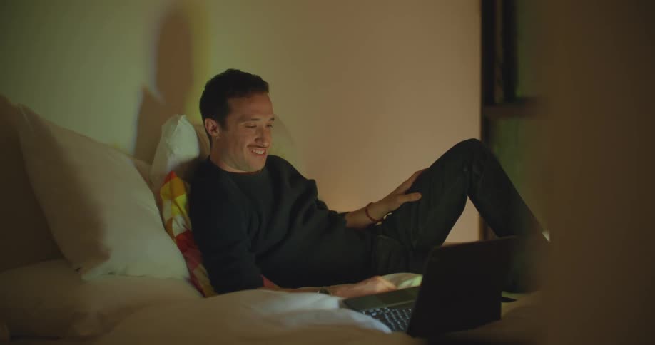 A man relaxes in bed watching a laptop screen and laughing