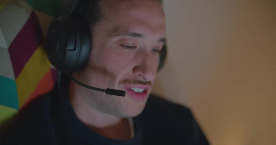 A young man sits on a couch and plays a video game as he talks in to a headset