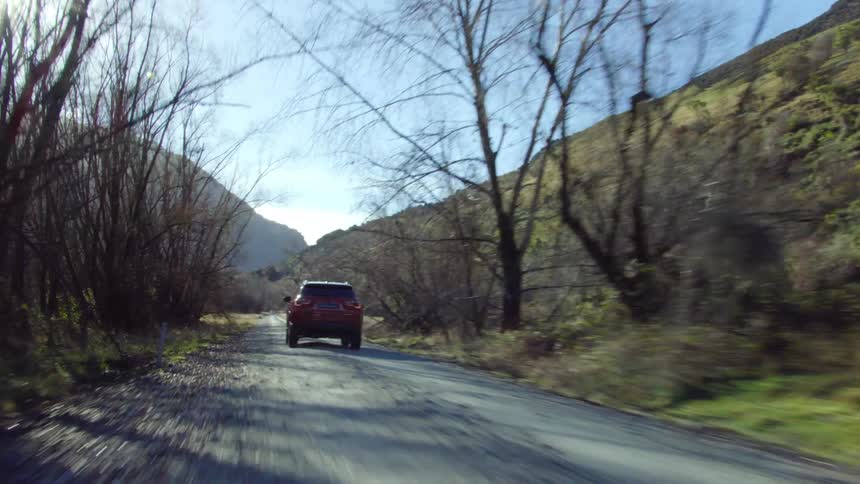 The sun is shining brightly on a red car as it travels on a road towards a mountain