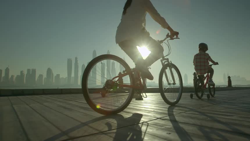 A woman and young boy bicycle across a boardwalk with the city in the background