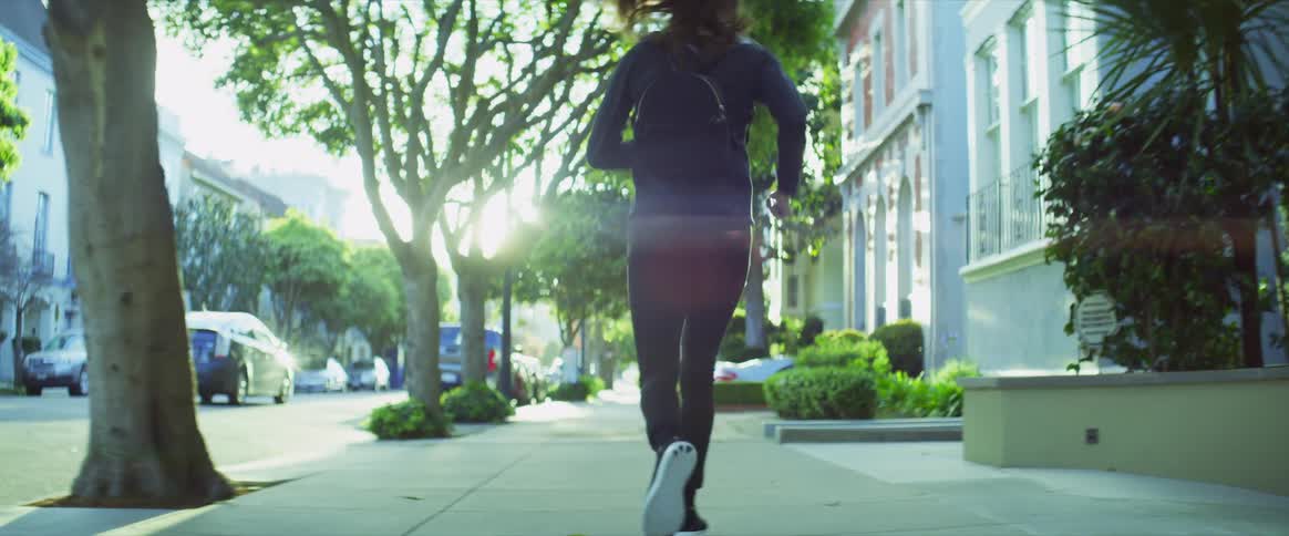 A woman wearing a backpack jogs down a sidewalk with trees and flowers on a bright sunny day