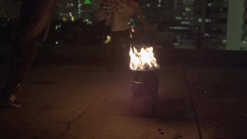 Teenagers riding skateboards around a barrel on flames on the roof of a building at night