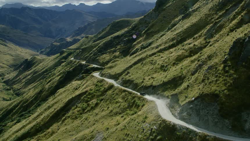 A vehicle travels along a winding mountain pass on the slope of a mountain covered in greenery