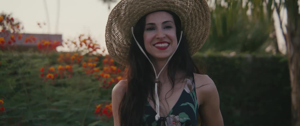 A woman with a hat laughing with a hedge flowers the sky and clouds in the background