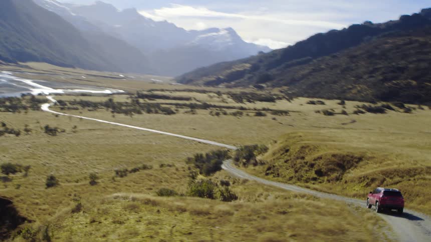 Vehicle travels on the road near the river and the fields with mountains nearby