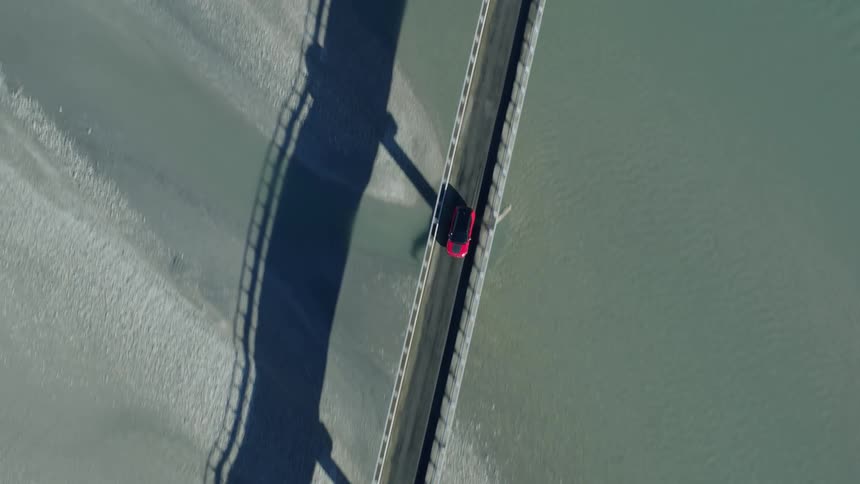 Red vehicle crosses long narrow bridge over blue gray water between muddy shores strewn with rocks