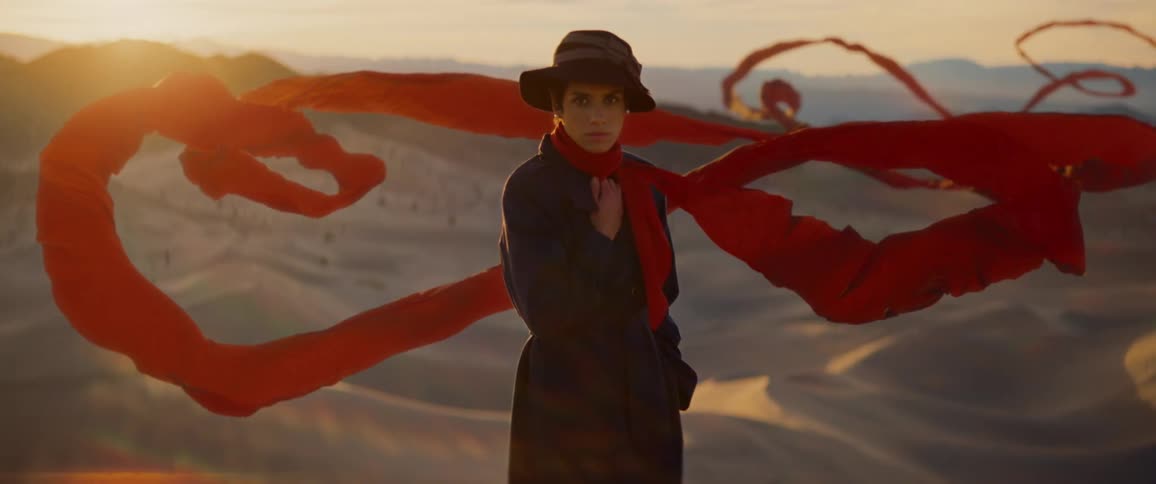 A woman wears a long trench coat as she stands in the desert and her long red scarf floats around her