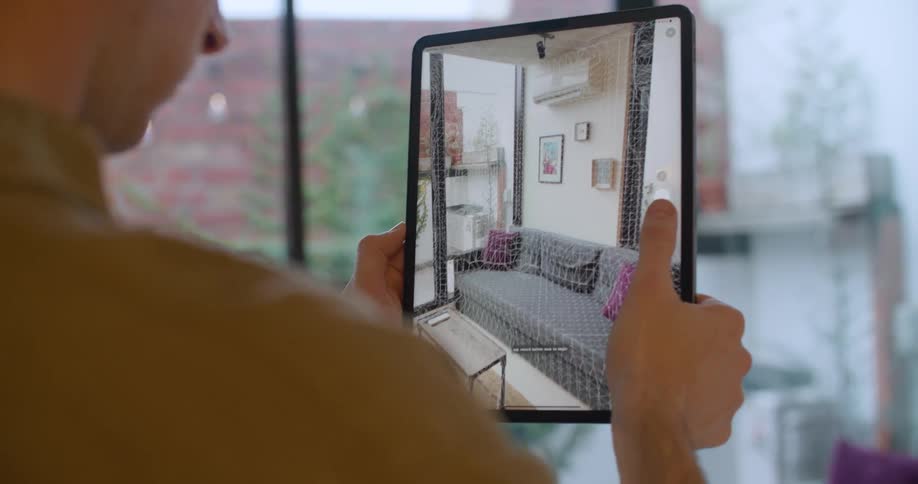 A man holds up a tablet and films the interior decor of an apartment including the deck and neighboring building