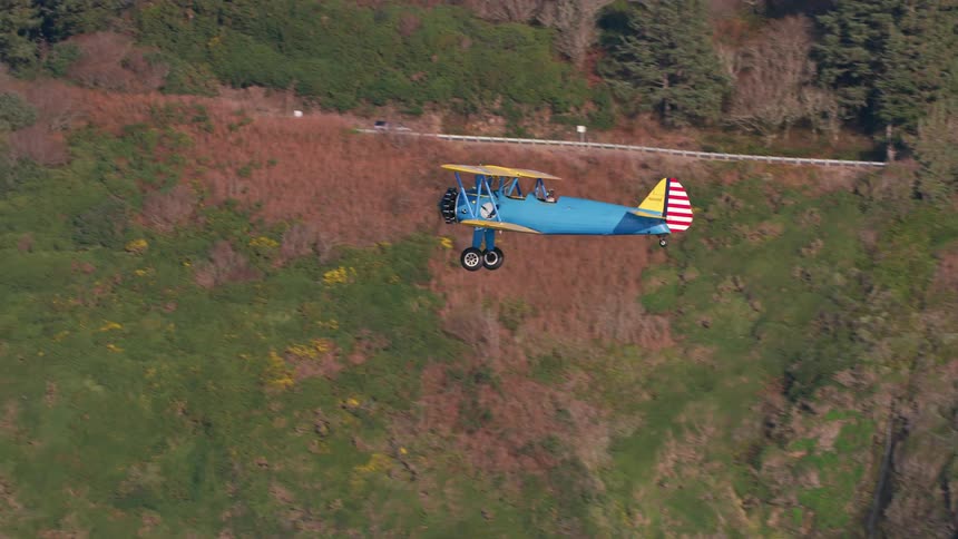 Blue biplane flies near the tree covered hills