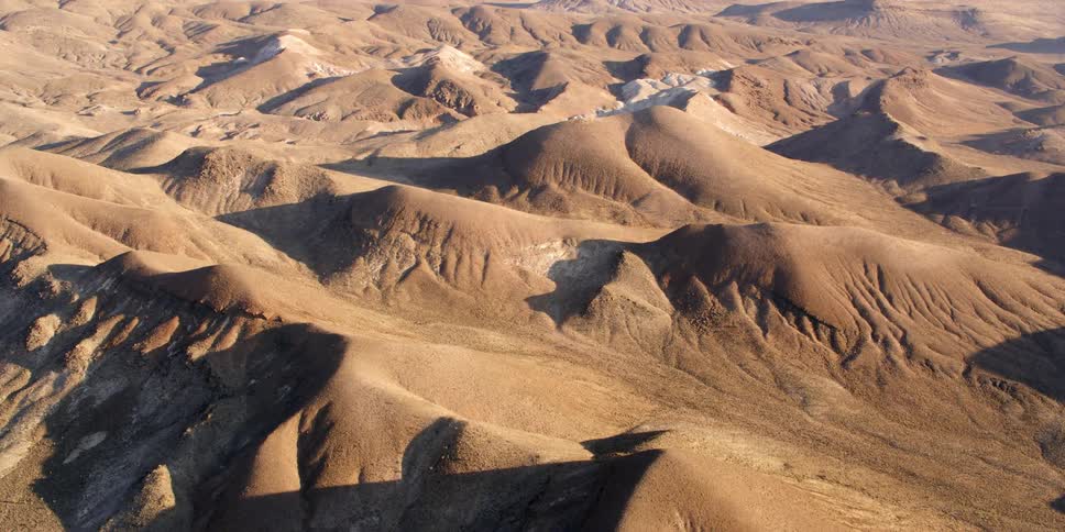 Bare sand and dirt cover hilly and mountainous terrain cast into light and shadow by the sun