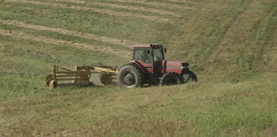 A person drives a farm tractor over a large field while towing a harvesting device behind it