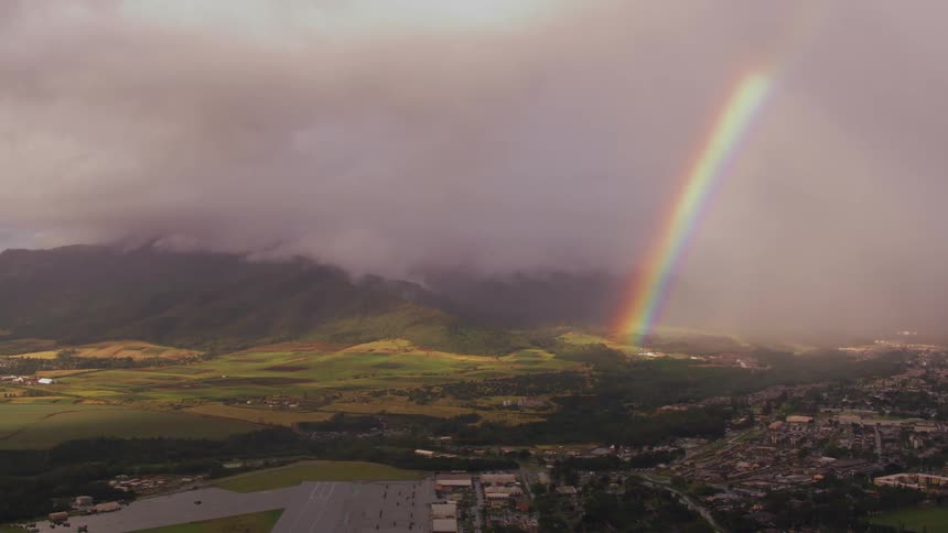 A rainbow is visible through thick clouds as it touches down between a field and a neighborhood